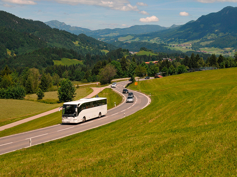 Moderner Reisebus fährt auf einer Bergstraße durch eine alpine Landschaft bei sonnigem Wetter und guter Sicht.